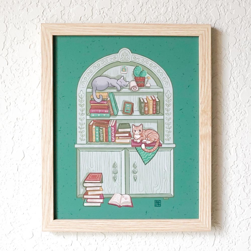 A framed print of a cute gray bookcase filled with books and trinkets with 2 cats lying on it. Background is green.