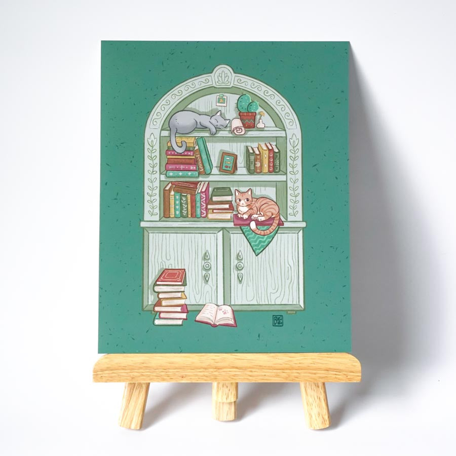 An unframed print on a easel of a cute gray bookcase filled with books and trinkets with 2 cats lying on it. Background is green.