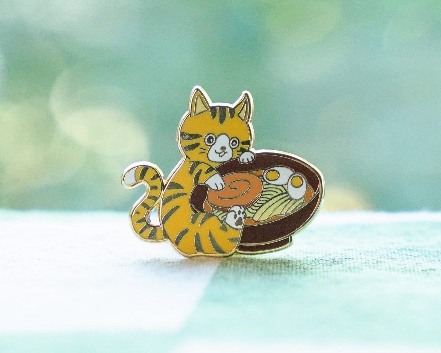 Enamel pin of a cat with a bowl of ramen on a blurred green background