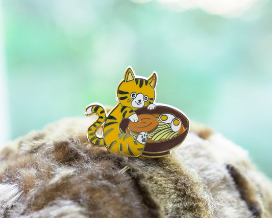 Enamel pin of a cartoon cat with a bowl on a textured surface with a blurred green background