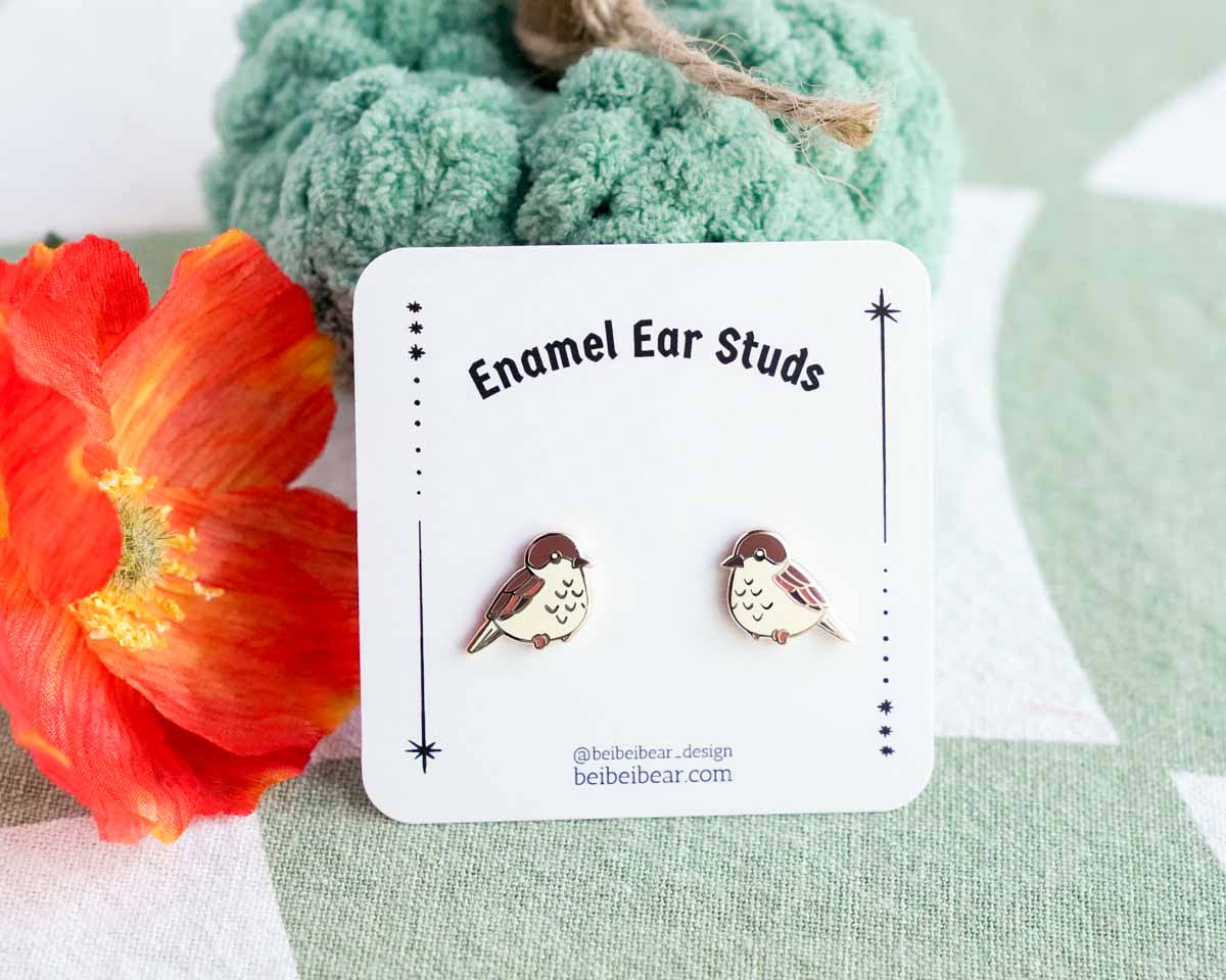 Bird-shaped enamel ear studs on a branded card with a floral background