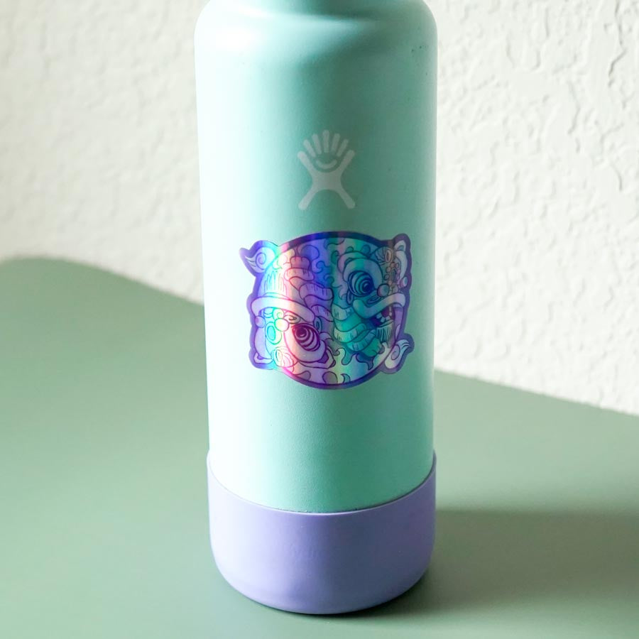 Turquoise and purple water bottle with a holographic yin yang lion dance design on a light green surface.